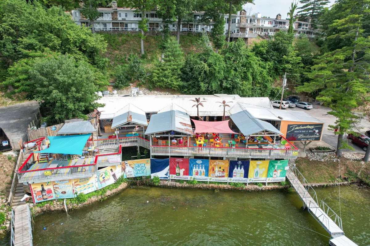 This exterior image captures a vibrant deck area overlooking the water, with colorful awnings and lively signage. Guests can be seen enjoying outdoor dining and social gatherings, set against the backdrop of a picturesque lake at sunset. The atmosphere is festive, hinting at themed events and local entertainment.