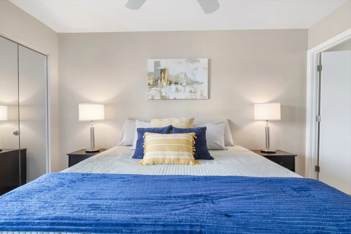 A cozy and serene bedroom featuring a bed with blue bedding and a yellow striped pillow, flanked by two bedside lamps. Abstract wall art and ample natural light contribute to the tranquil, refreshing ambiance.