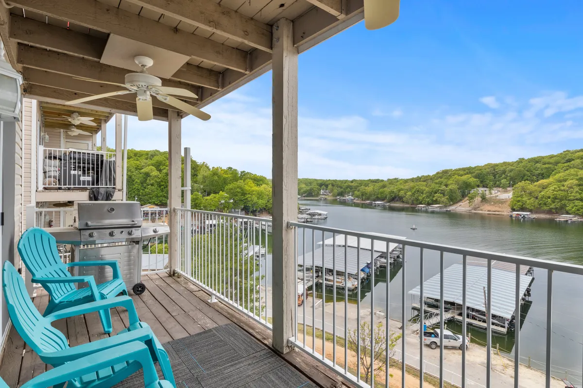 The image showcases a serene deck with inviting blue Adirondack chairs overlooking the sparkling lake at Lake Ozark. A grill sits nearby, perfect for outdoor cooking, while the tranquil water and lush shoreline set a peaceful, relaxing mood. The view promises breathtaking sunsets and a direct connection to the natural beauty of the cove, suggesting easy access for boating or fishing.