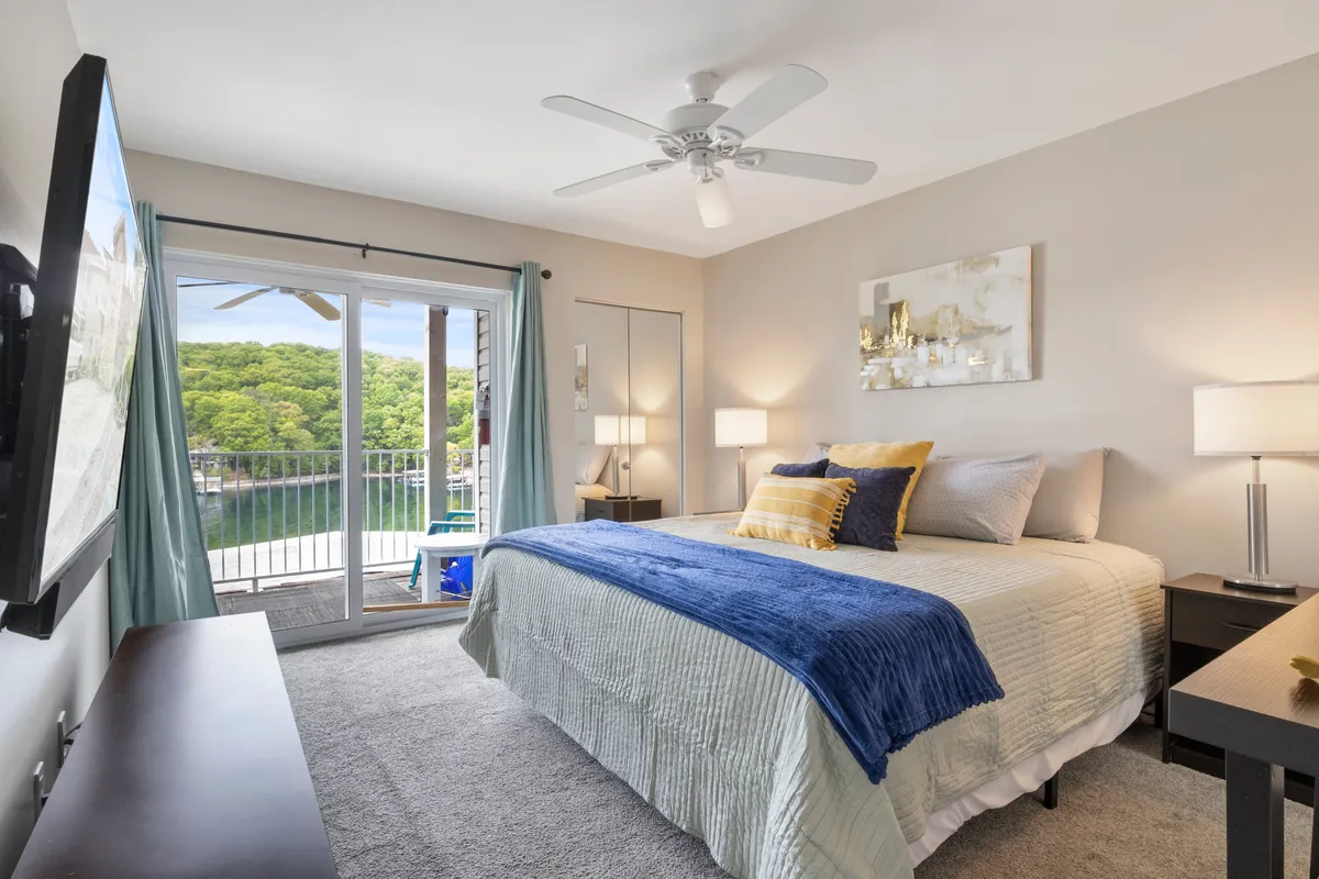 This image captures a serene bedroom featuring a bed with a blue throw blanket, a large TV mounted on the wall, and a sliding glass door leading to a balcony with views of nature. The space is filled with soft natural light and tranquil decor, creating an inviting atmosphere ideal for relaxation and restful sleep.