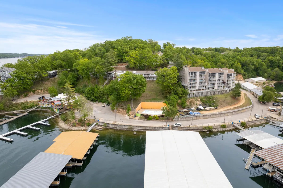 A picturesque view showcasing the calm lake, lush green hillside, a residential building, and docks extending into the water. The image exudes a peaceful lakeside atmosphere with plenty of open space and spots for families to gather, fish, picnic, or relax by the water's edge.